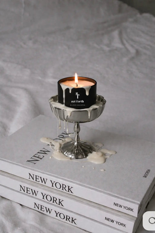 Lit Gilded Whisper candle in matte black tin on stacked New York magazines with wax dripping.